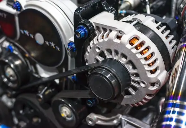 Automotive Repair & Tire Shop Oakville, ON - Sil’s Auto Care Centre Close-up of a car engine with a visible alternator, belts, and metal components. The alternator’s copper coils and pulley are prominently shown, with other engine parts and wires surrounding it. | Call Us Today At +1 (905) 823-2626