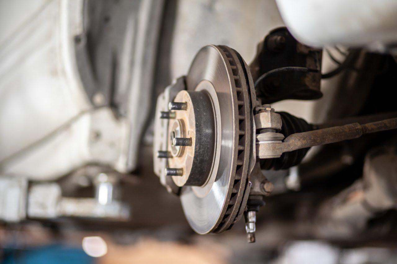 Close-up of a vehicles disc brake system, showcasing a ventilated brake disc, caliper, and wheel hub assembly. Expertly crafted for perfect alignment & steering by The Mufflerman, the components sit beneath the cars wheel well against a blurred white background.