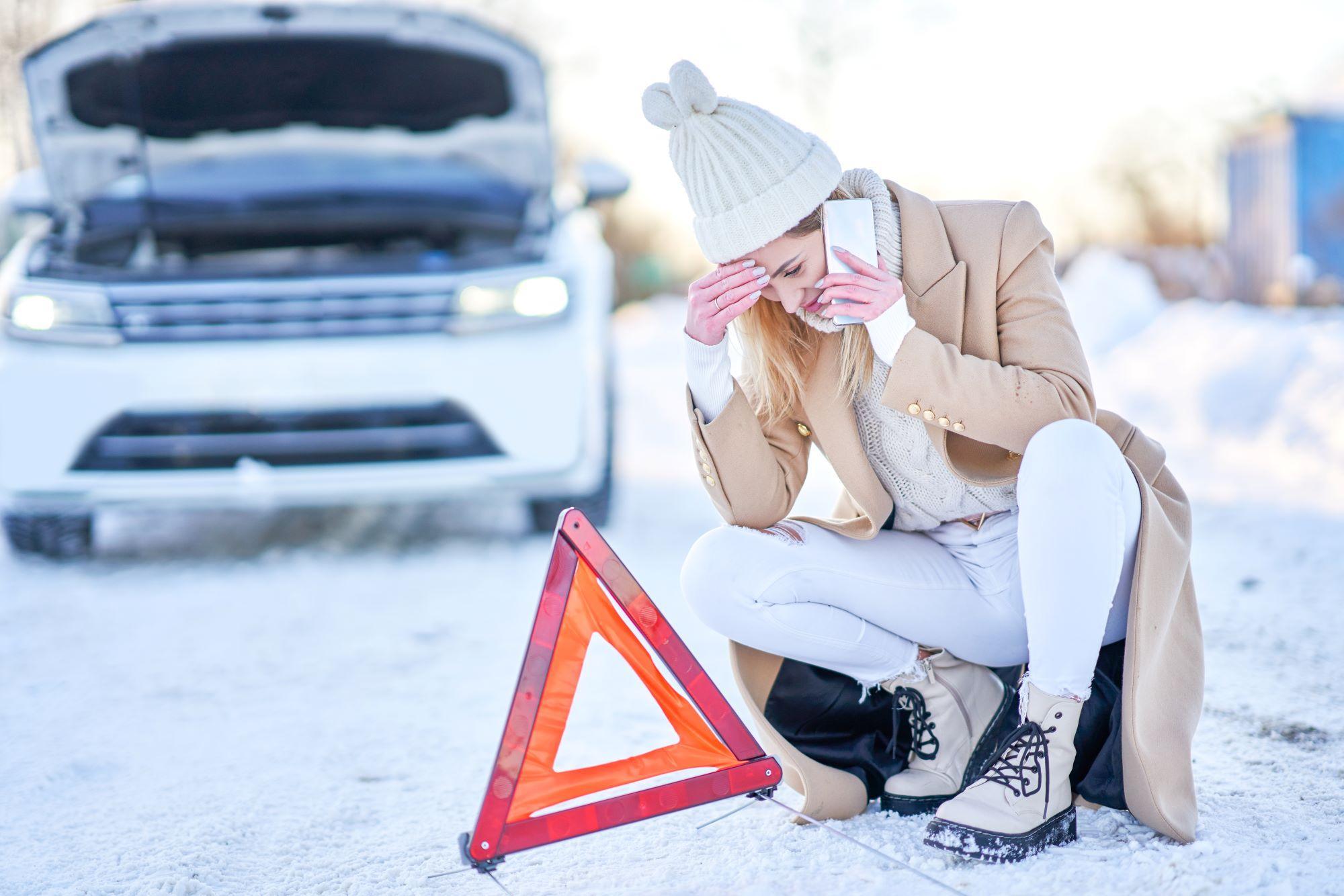 Automotive Repair & Tire Shop Oakville, ON - Sil’s Auto Care Centre A woman in winter clothing sits by the roadside with her head in her hand, talking on the phone. A warning triangle is in front of her, and a car with its bonnet up is parked behind her on a snowy road—perfect reminder for winter car maintenance tips. | Call Us Today At +1 (905) 823-2626