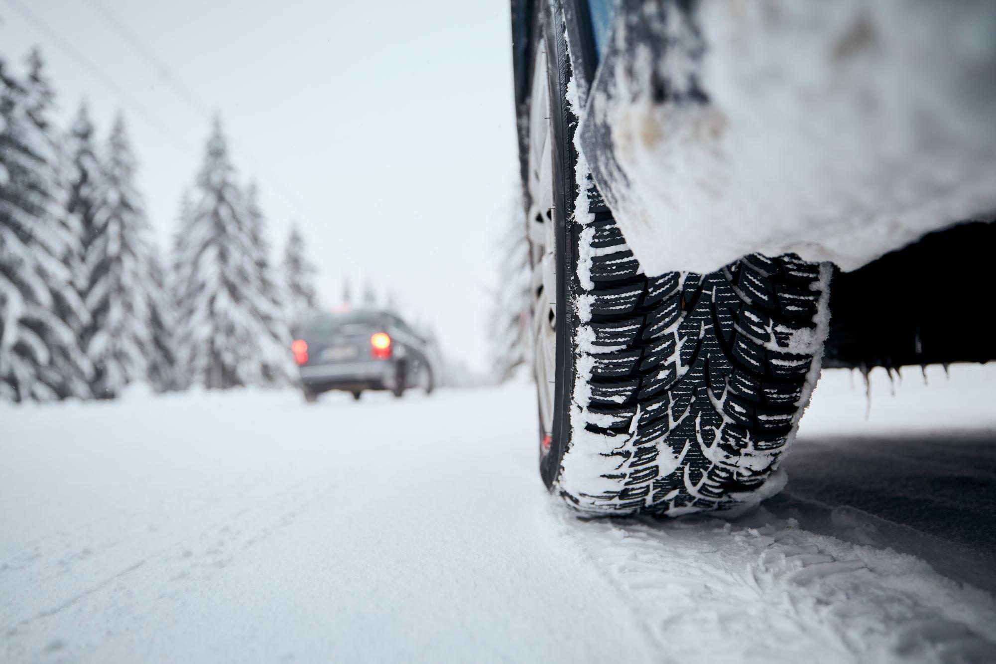 Automotive Repair & Tire Shop Oakville, ON - Sil’s Auto Care Centre Close-up of a car tyre driving on a snow-covered road in a snowy forest. Another vehicle is visible ahead, and tall trees coated with snow line the sides of the road—a perfect scene for showcasing Winter Tyres 101 in action. | Call Us Today At +1 (905) 823-2626