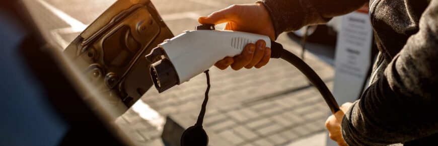 Automotive Repair & Tire Shop Oakville, ON - Sil’s Auto Care Centre A Hybrid Car Mechanic holds a charging plug, preparing to connect it to an electric vehicle at a charging station in an outdoor car park. | Call Us Today At +1 (905) 823-2626
