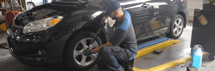 Automotive Repair & Tire Shop Oakville, ON - Sil’s Auto Care Centre A mechanic in uniform is using a tool to work on the front tyre of a black car at a Car Repair Mississauga shop. The car’s bonnet is open, with various equipment and supplies visible throughout the bustling auto repair shop. | Call Us Today At +1 (905) 823-2626