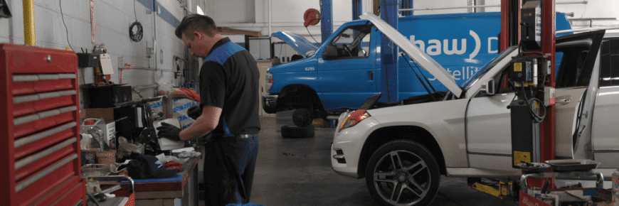 Automotive Repair & Tire Shop Oakville, ON - Sil’s Auto Care Centre A mechanic examines paperwork at a workbench in a car repair shop. Nearby, a white SUV and a blue van with open bonnets are being serviced—showcasing the busy atmosphere of car repair near me. Tools and equipment are visible throughout the garage. | Call Us Today At +1 (905) 823-2626