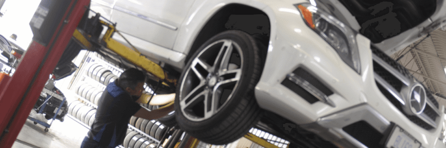 Automotive Repair & Tire Shop Oakville, ON - Sil’s Auto Care Centre A mechanic works underneath a white car lifted on a hydraulic lift in a car repair Oakville workshop, with stacks of tyres visible in the background. | Call Us Today At +1 (905) 823-2626
