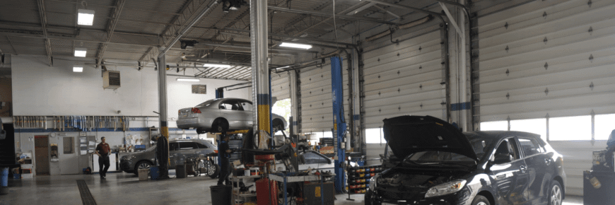 Automotive Repair & Tire Shop Oakville, ON - Sil’s Auto Care Centre An auto repair shop interior with cars on lifts, one car with an open bonnet, tools and equipment scattered around, and a car mechanic near me working near the back; large garage doors line the right side. | Call Us Today At +1 (905) 823-2626