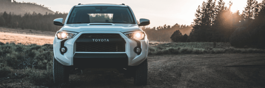 Automotive Repair & Tire Shop Oakville, ON - Sil’s Auto Care Centre A white Toyota SUV is parked on a dirt track at sunset, its headlights shining. Trees and grassy hills are in the background under a partly cloudy sky—a perfect scene for those searching "Toyota Mechanic Near Me" after an evening drive. | Call Us Today At +1 (905) 823-2626