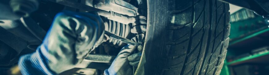 Automotive Repair & Tire Shop Oakville, ON - Sil’s Auto Care Centre A mechanic wearing gloves works on a car’s suspension or steering system, using a tool near the wheel and tyre. | Call Us Today At +1 (905) 823-2626