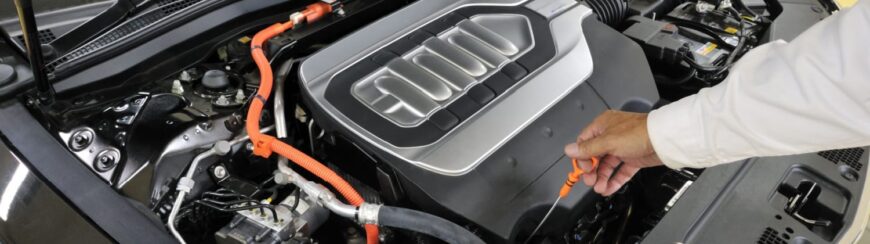 Automotive Repair & Tire Shop Oakville, ON - Sil’s Auto Care Centre A person’s hand pulling the orange dipstick from the engine compartment of an electric or hybrid vehicle, revealing part of the engine and various cables under the bonnet. | Call Us Today At +1 (905) 823-2626