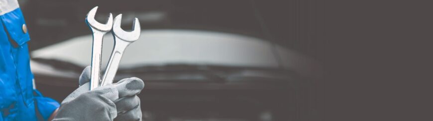 Automotive Repair & Tire Shop Oakville, ON - Sil’s Auto Care Centre A person wearing grey gloves and a blue uniform holds two spanners in front of a blurred car bonnet background, with ample empty space on the right side of the image. | Call Us Today At +1 (905) 823-2626