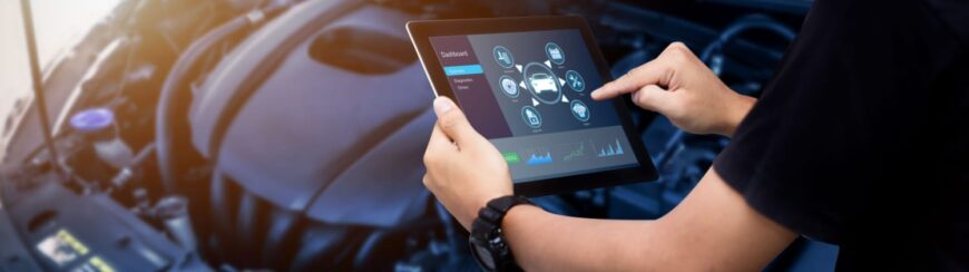 Automotive Repair & Tire Shop Oakville, ON - Sil’s Auto Care Centre A person holds a tablet displaying a car diagnostics interface in front of an open car engine, using the touchscreen to check vehicle data and performance metrics. | Call Us Today At +1 (905) 823-2626