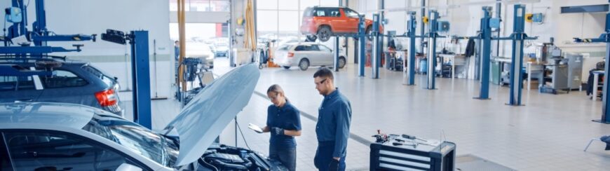 Automotive Repair & Tire Shop Oakville, ON - Sil’s Auto Care Centre Two auto mechanics, one holding a tablet, inspect a car with its bonnet open in a spacious, well-lit garage. Several cars are seen on lifts and technicians work in the background. | Call Us Today At +1 (905) 823-2626