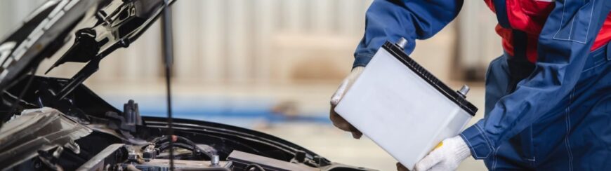 Automotive Repair & Tire Shop Oakville, ON - Sil’s Auto Care Centre A mechanic wearing gloves and a blue uniform holds a white car battery next to an open car bonnet, preparing to install or replace it in a garage setting. | Call Us Today At +1 (905) 823-2626