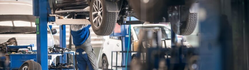 Automotive Repair & Tire Shop Oakville, ON - Sil’s Auto Care Centre A mechanic in blue overalls works underneath a car lifted on a hydraulic ramp in a brightly lit car repair workshop with multiple vehicles and equipment visible. | Call Us Today At +1 (905) 823-2626