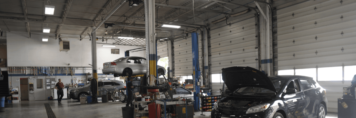 Automotive Repair & Tire Shop Oakville, ON - Sil’s Auto Care Centre An auto repair shop interior with cars on lifts, one car with an open bonnet, tools and equipment scattered around, and a car mechanic near me working near the back; large garage doors line the right side. | Call Us Today At +1 (905) 823-2626