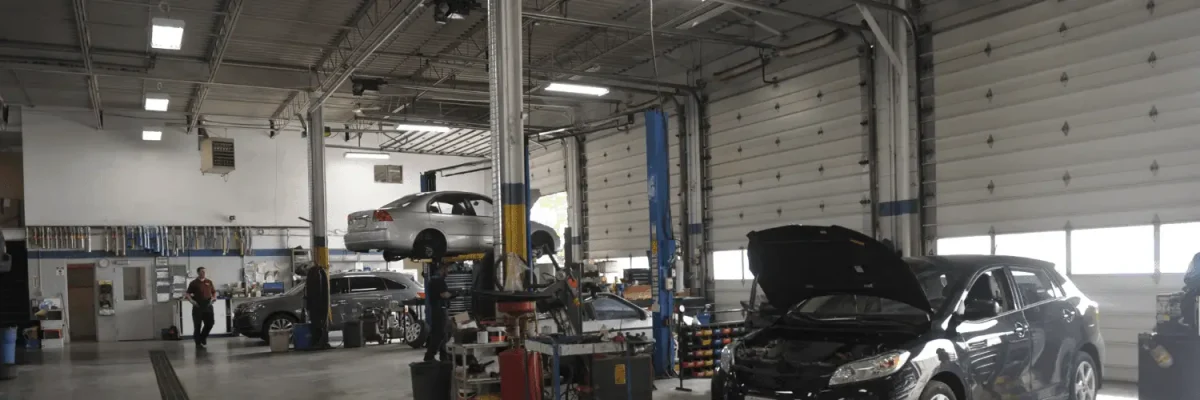 Automotive Repair & Tire Shop Oakville, ON - Sil’s Auto Care Centre An auto repair shop interior with cars on lifts, one car with an open bonnet, tools and equipment scattered around, and a car mechanic near me working near the back; large garage doors line the right side. | Call Us Today At +1 (905) 823-2626