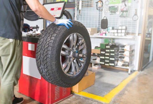 Automotive Repair & Tire Shop Oakville, ON - Sil’s Auto Care Centre A person wearing gloves is holding a large car tyre mounted on a wheel balancing machine inside a car repair shop with tools and parts in the background. | Call Us Today At +1 (905) 823-2626