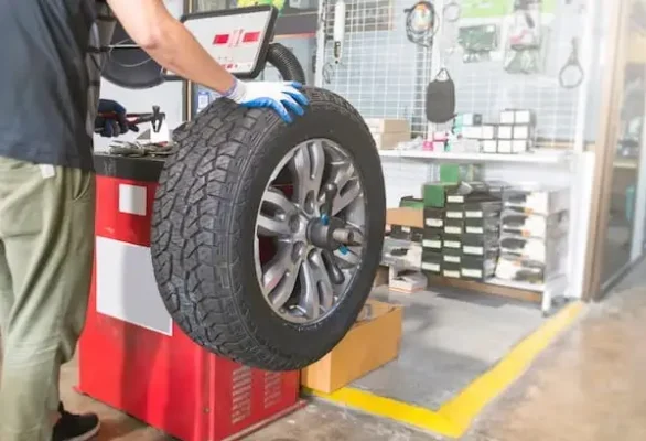 Automotive Repair & Tire Shop Oakville, ON - Sil’s Auto Care Centre A person wearing gloves is holding a large car tyre mounted on a wheel balancing machine inside a car repair shop with tools and parts in the background. | Call Us Today At +1 (905) 823-2626