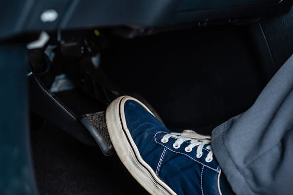 Automotive Repair & Tire Shop Oakville, ON - Sil’s Auto Care Centre A person wearing blue trainers presses down a car pedal, likely the brake or clutch, inside a vehicle. | Call Us Today At +1 (905) 823-2626