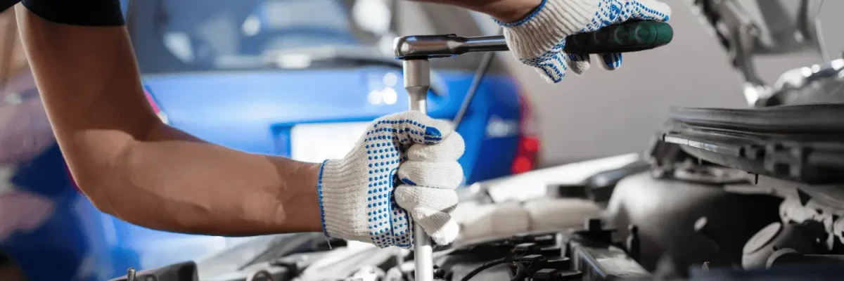 Automotive Repair & Tire Shop Oakville, ON - Sil’s Auto Care Centre A person wearing white work gloves uses a ratchet spanner to work on a car engine, with a blue car in the blurred background—capturing the attention of anyone searching for "Car Repair Near Me. | Call Us Today At +1 (905) 823-2626