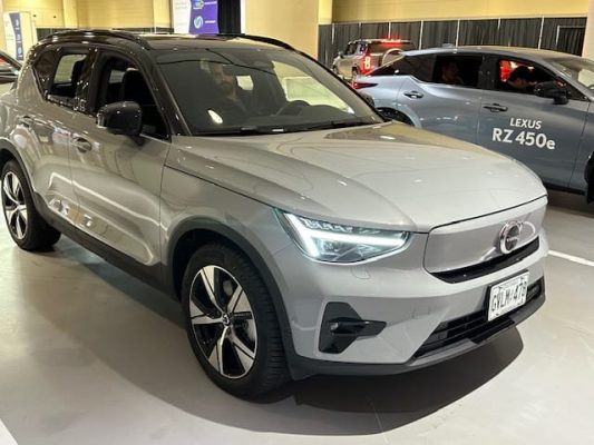 Automotive Repair & Tire Shop Oakville, ON - Sil’s Auto Care Centre A silver Volvo electric SUV is displayed indoors under bright lights, with a Lexus RZ 450e parked nearby. The car has sleek headlamps, black accents, and a front number plate. | Call Us Today At +1 (905) 823-2626