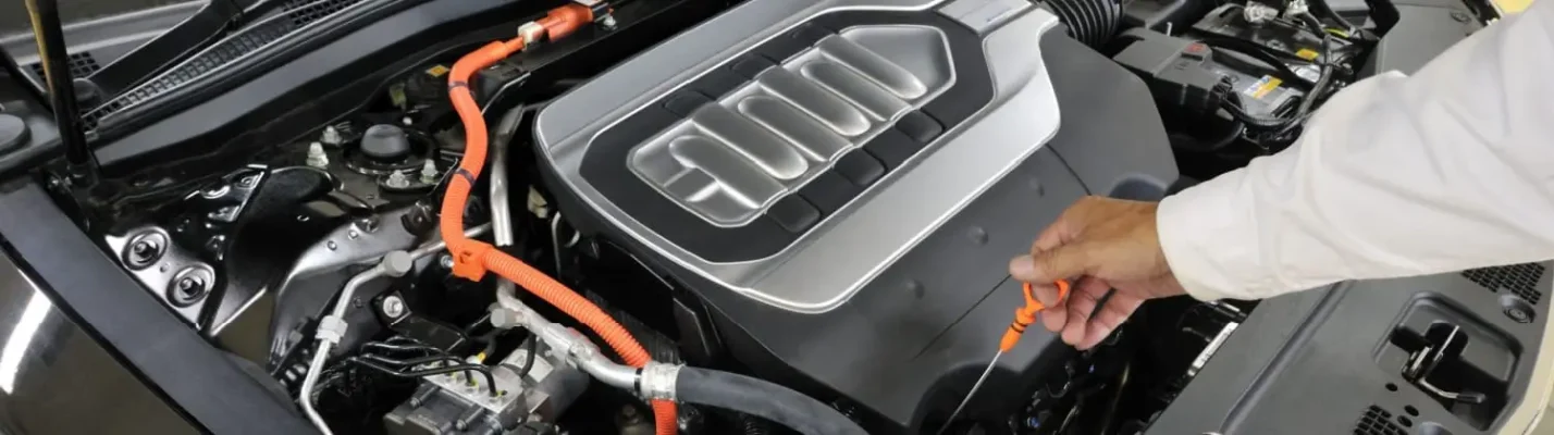 Automotive Repair & Tire Shop Oakville, ON - Sil’s Auto Care Centre A person’s hand pulling the orange dipstick from the engine compartment of an electric or hybrid vehicle, revealing part of the engine and various cables under the bonnet. | Call Us Today At +1 (905) 823-2626