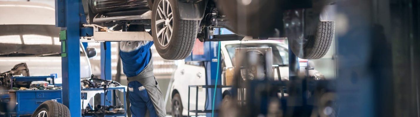 Automotive Repair & Tire Shop Oakville, ON - Sil’s Auto Care Centre A mechanic in blue overalls works underneath a car lifted on a hydraulic ramp in a brightly lit car repair workshop with multiple vehicles and equipment visible. | Call Us Today At +1 (905) 823-2626