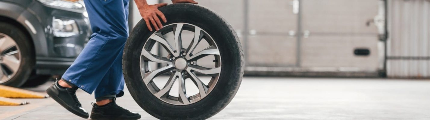 Automotive Repair & Tire Shop Oakville, ON - Sil’s Auto Care Centre A person in blue trousers and black shoes rolls a car tyre across a garage floor, with part of a vehicle visible in the background. | Call Us Today At +1 (905) 823-2626
