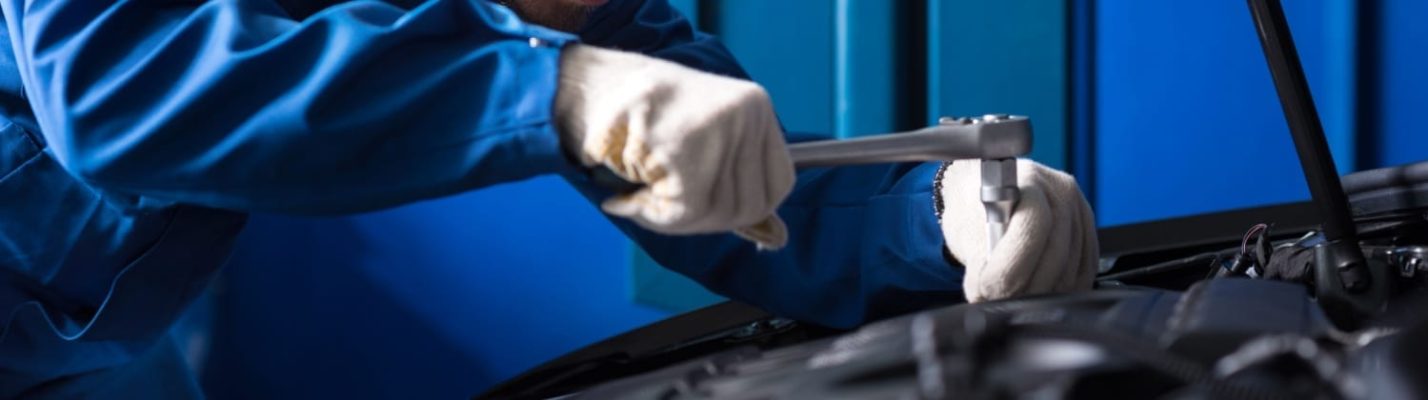 Automotive Repair & Tire Shop Oakville, ON - Sil’s Auto Care Centre A mechanic in blue overalls and white gloves uses a spanner to work on a car engine in a garage. | Call Us Today At +1 (905) 823-2626