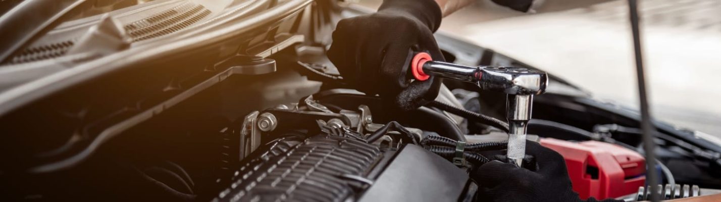Automotive Repair & Tire Shop Oakville, ON - Sil’s Auto Care Centre A close-up of a mechanic’s gloved hands using a ratchet spanner to work on the engine of a car. Various engine components and tools are visible under the open bonnet. | Call Us Today At +1 (905) 823-2626