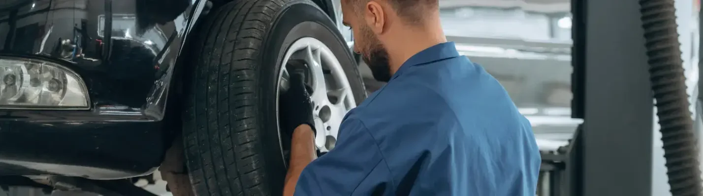 Automotive Repair & Tire Shop Oakville, ON - Sil’s Auto Care Centre A mechanic in a blue uniform works on a car’s front tyre in a garage, with the car lifted on a hydraulic lift. | Call Us Today At +1 (905) 823-2626