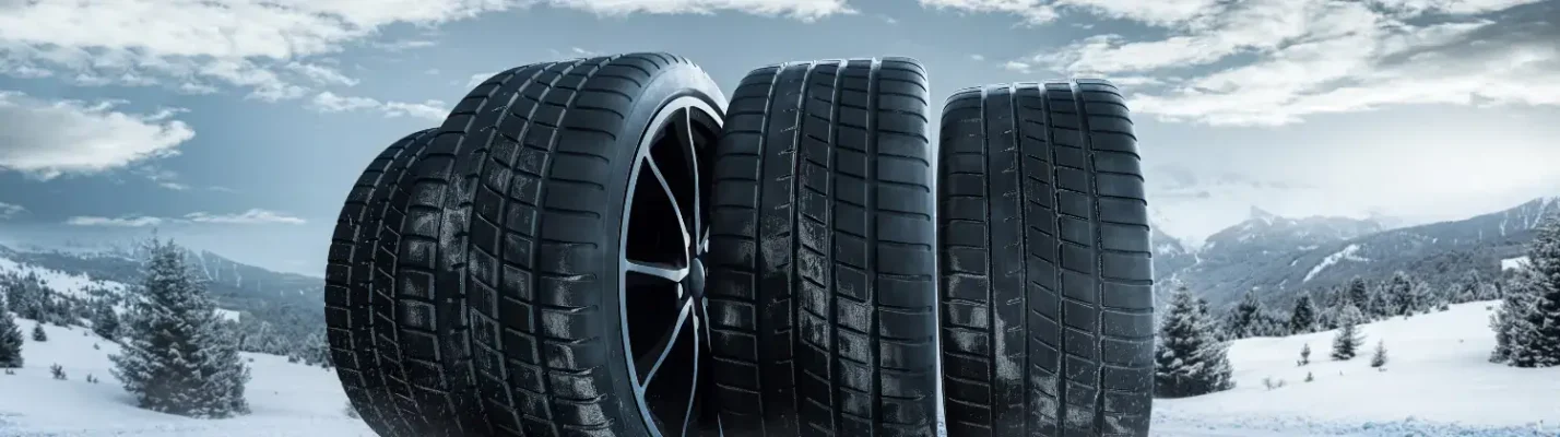 Automotive Repair & Tire Shop Oakville, ON - Sil’s Auto Care Centre Four car tyres with deep treads are standing upright on a snowy landscape with snow-covered trees and mountains in the background under a partly cloudy sky. | Call Us Today At +1 (905) 823-2626