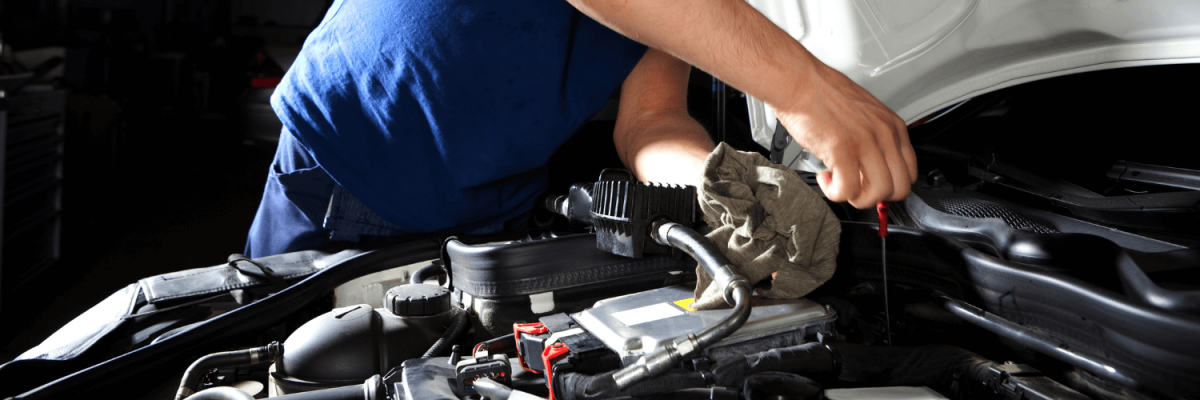 Automotive Repair & Tire Shop Oakville, ON - Sil’s Auto Care Centre A person in a blue shirt checks the oil level of a car engine under the bonnet, holding a cloth and dipstick in a well-lit garage—just what you'd expect from a car mechanic near me. | Call Us Today At +1 (905) 823-2626