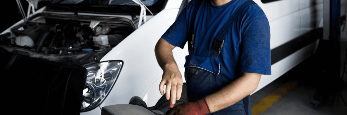 Automotive Repair & Tire Shop Oakville, ON - Sil’s Auto Care Centre A mechanic in blue overalls and red gloves works on a vehicle part in a garage, with a white van and its open bonnet visible in the background—perfect for anyone searching for Car Repair Garages Near Me. | Call Us Today At +1 (905) 823-2626